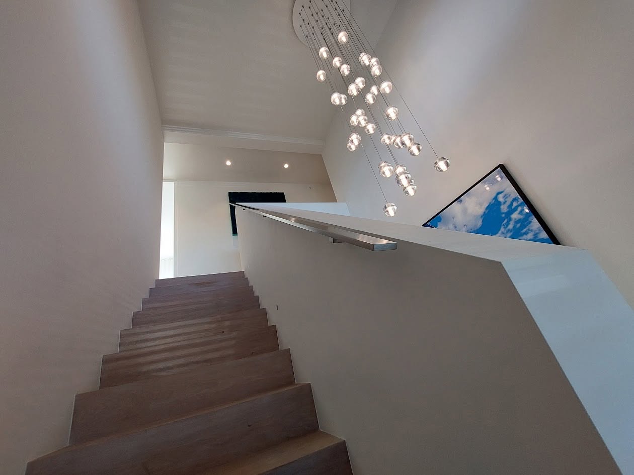 Houten trap met strakke stalen leuning onder een vide met hanglamp